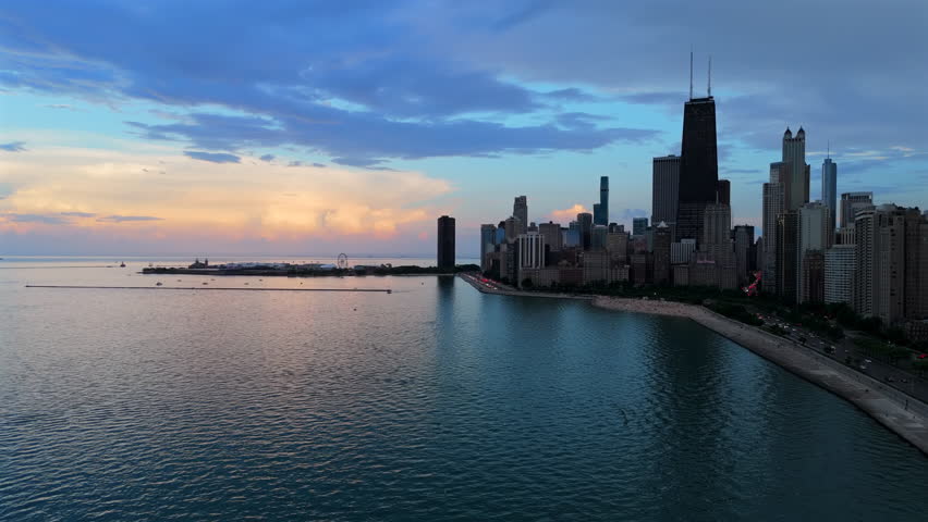 Aerial drone footage captures the stunning Chicago skyline along Lake Michigan during sunset. Tall skyscrapers reflect in calm waters as colorful clouds paint the sky in pink and blue hues.