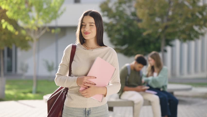 Portrait of a pretty young woman holding books and smiling at the camera. College student standing on campus with classmates in the background