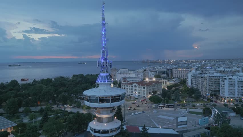 Dramatic aerial view of the illuminated OTE Tower in Thessaloniki at blue hour featuring a spectacular lightning strike in the stormy sky over the sea.