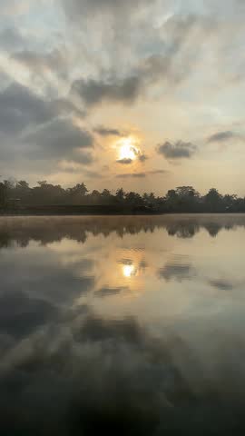 Serene sunrise over a calm lake with morning mist and cloud reflections. soft sunrise breaking through dramatic clouds.