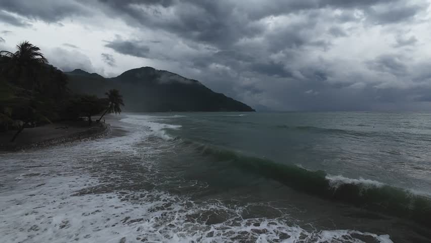 Storm waves approaching tropical coastline. Aerial drone view of rolling ocean swell under heavy clouds with mountain backdrop, creating dramatic seascape with rough water and storm atmosphere.