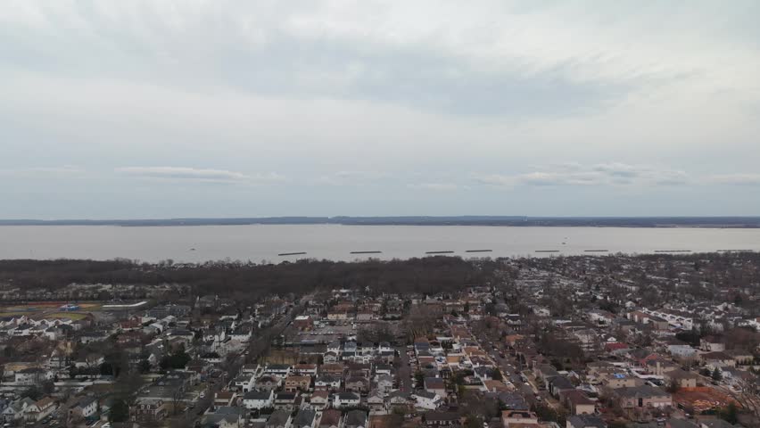 High altitude aerial perspective slowly flying over a suburban town with houses and leafless trees during a cold, cloudy winter day, approaching the expansive waterfront of a bay