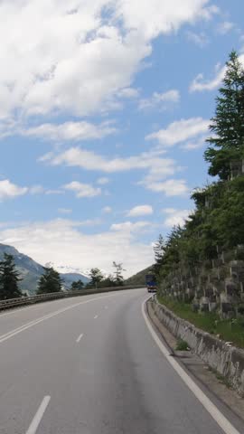 Vertical POV shot of a heavy truck driving uphill on a winding mountain road in the French Alps. Scenic highway with snow-capped peaks and green slopes under a blue sky