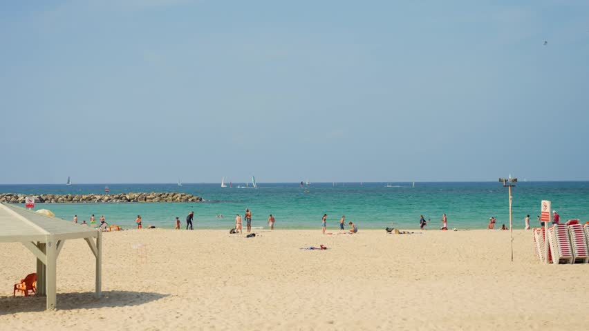 Wide shot of Tel Aviv beach during summer day as people swim and suntan on sand. Locals and tourists enjoy sunny hot day outdoors playing by Mediterranean Sea in Israel. Blue water and sails