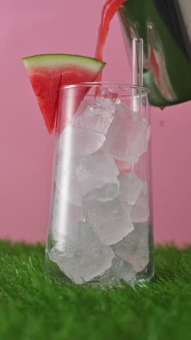 Fresh watermelon drink pours into iced glass with garnish and straw on pink background in bright summer refreshment scene