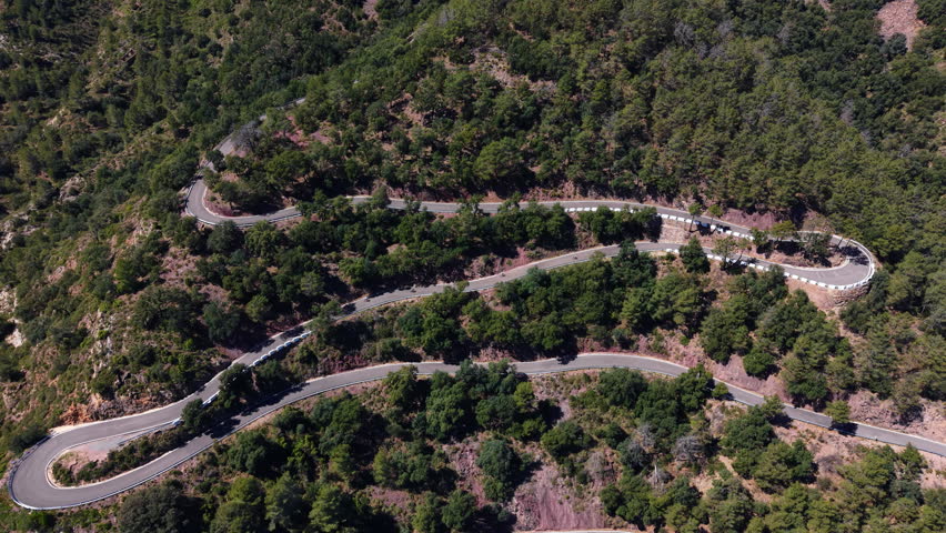Aerial view of cyclists descending a serpentine mountain road with sharp hairpin bends through dense green forest in Sierra de Espadan, Castellon, Spain