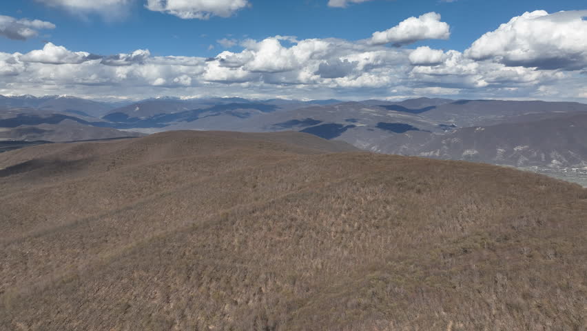 Aerial view of a dense mountain forest with trees in early spring colors under a clear blue sky with puffy white clouds
