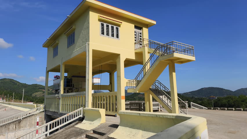 Dam control and room building stand on elevated pillars above the infrastructure. Bright yellow structure features exterior stairs and metal railings. This dam control unit operates from room building