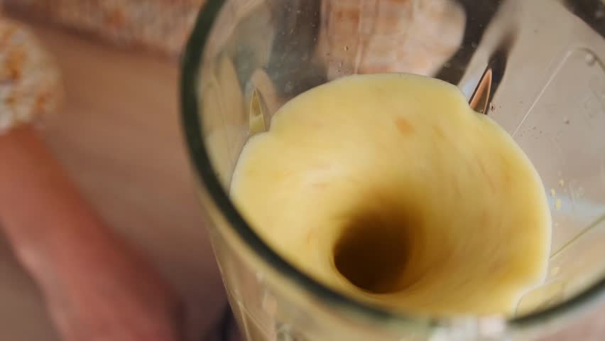 Close-up, young woman in a textured brown sweater preparing a fresh smoothie in a blender in a modern kitchen. Thick yellow smoothie is being poured into a glass. Process of making a healthy drink