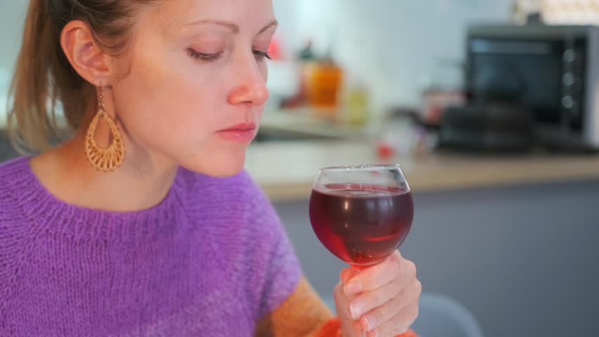 Young woman in a purple sweater enjoying a glass of red wine, thoughtfully holding and sipping from the wine glass, with her eyes closed, moment of relaxation and enjoyment, at home kitchen. Close-up.