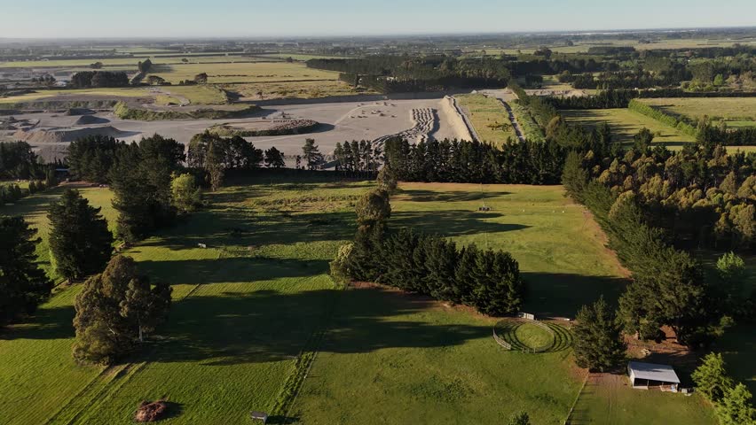 Aerial view of rural landscape near Sheffield outside Christchurch, New Zealand, with farmland, tree-lined roads and distant mountains glowing in warm golden sunset light. Panning.