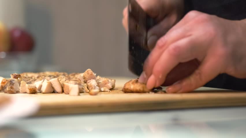 A close-up view of hands cutting grilled chicken breast on a wooden board. The juicy meat is sliced with precision using a kitchen knife. Cooking process concept.
