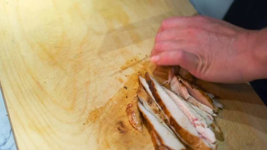 Close-up of a person slicing grilled chicken breast into small pieces on a wooden cutting board. Juicy meat with a crispy golden crust. Cooking preparation, hands, knife, and textures of the food.