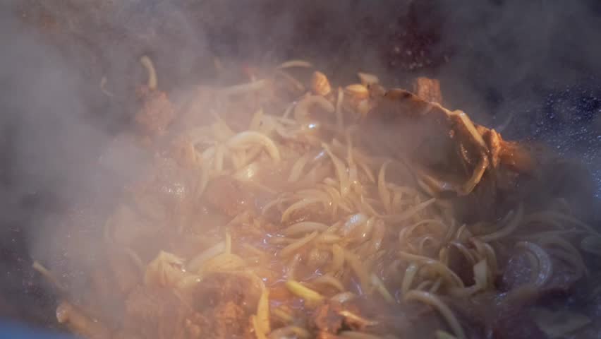 Traditional pilaf preparation with meat and onions being stir-fried in a hot cauldron. Steam rises as ingredients are mixed. Rich colors and textures of sizzling food captured in close-up.