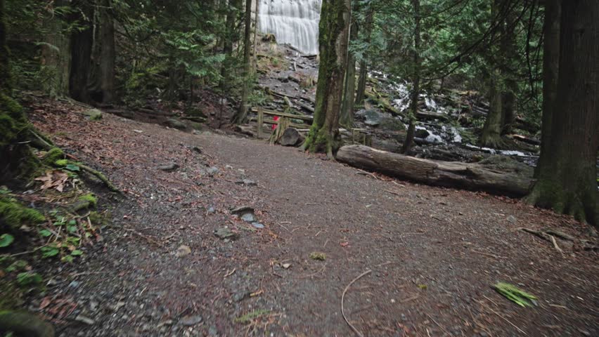 A smooth tracking shot pushes forward on a forest trail toward a wooden barrier and a red warning sign. The dense green woods frame the stunning cascading water of Bridal Veil Falls in the distance
