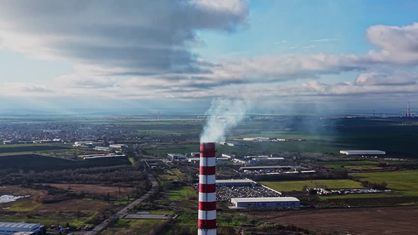 Smoke rises from a tall factory smokestack over an industrial area. Fields and buildings extend into the distance under a partly cloudy sky. This scene shows pollution and industry.