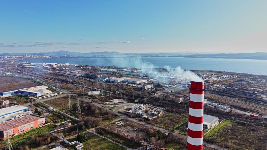 A power plant stands by the water with smoke rising from its chimney. Industrial buildings and structures can be seen nearby along with a clear sky and distant hills.