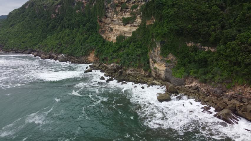 Aerial view of rugged coastal cliffs covered in lush greenery with ocean waves crashing against rocky shoreline, creating dramatic natural landscape with textured rock formations.