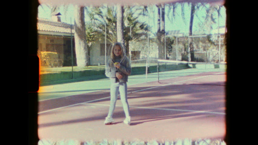 Authentic vintage home movie from the 1970s. A young blonde girl smiles while holding a tennis racket and ball on a sunny court. Nostalgic, grainy Super 8 film look with light leaks.