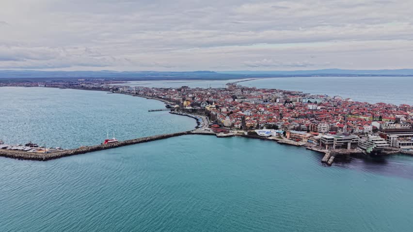 Aerial view showcases a town along the coast with many buildings facing the water. A pier extends into the sea, and boats are visible nearby. Clouds cover the sky above.