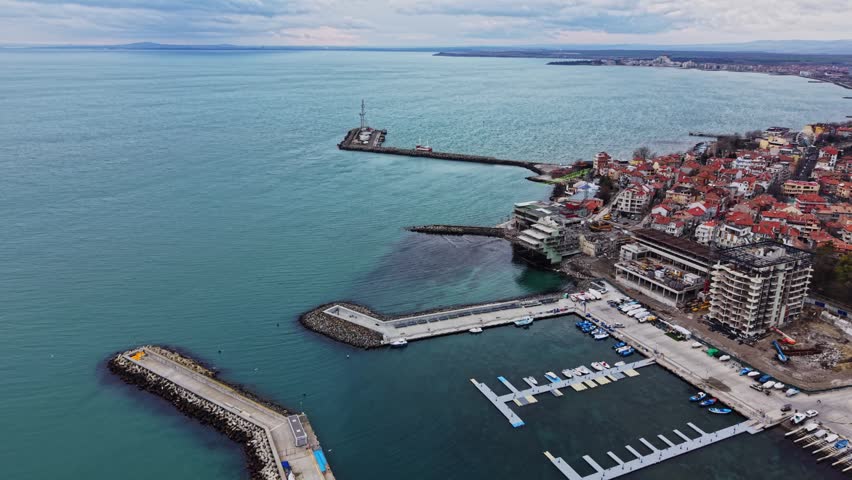 The scene shows a coastal area with a harbor and various boats docked. Buildings are lined along the waterfront under a cloudy sky. Water stretches to the horizon.