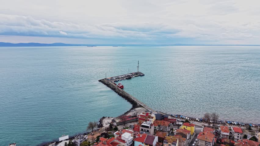 A clear view of the water stretches out from a high point, with boats anchored in the harbor. Houses with red rooftops cover the land near the shore, under a cloudy sky.
