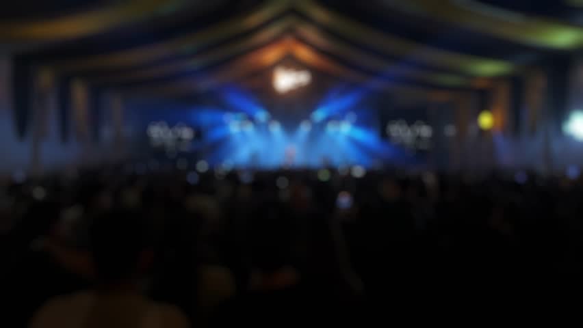 Video of silhouette people having fun at a rock concert with a band performing on a blurry lit stage. Energetic live music event footage.