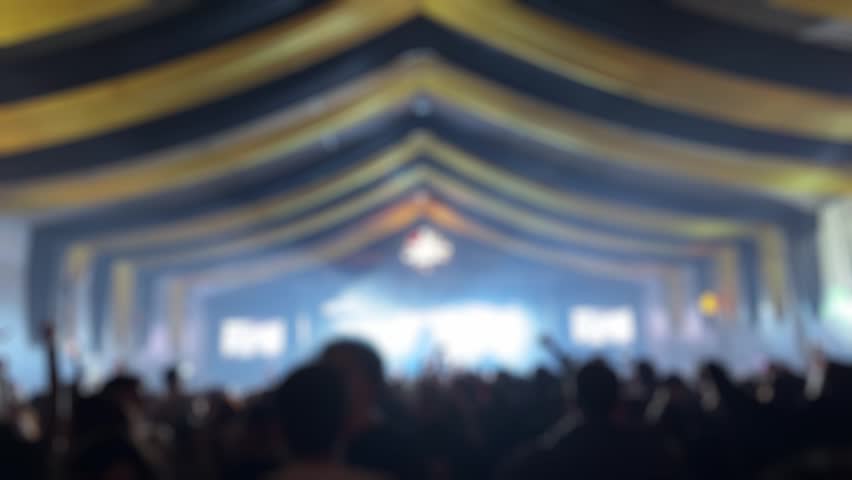 Video of silhouette people having fun at a rock concert with a band performing on a blurry lit stage. Energetic live music event footage.