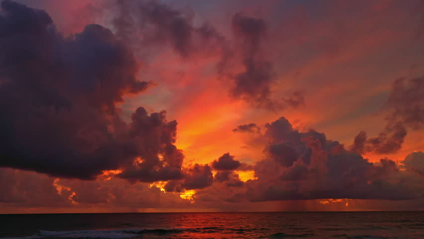 A stunning coastal landscape featuring a vibrant purple and orange sunset over the ocean, with dramatic clouds illuminated by the last rays of sunlight. Gentle waves roll toward the sandy shore