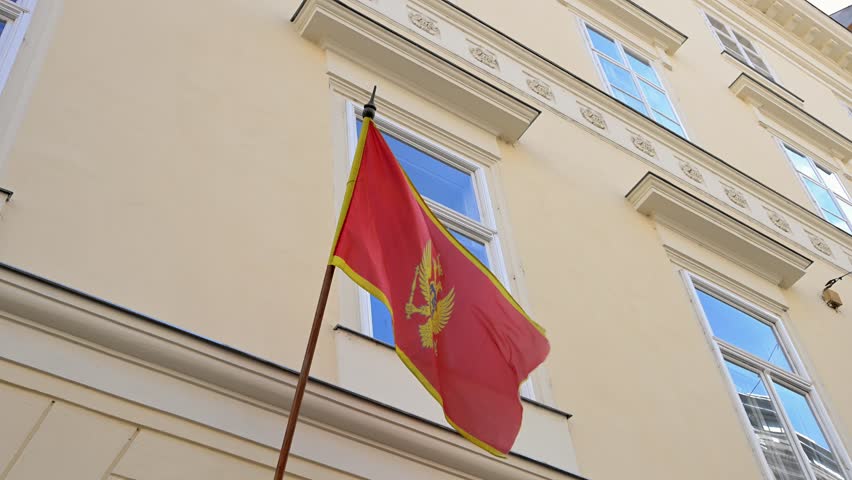 A low-angle shot shows the red national flag of Montenegro with its golden coat of arms hanging from a wooden pole against a cream-colored classical building. The architectural background features several windows with white frames and decorative moldings.