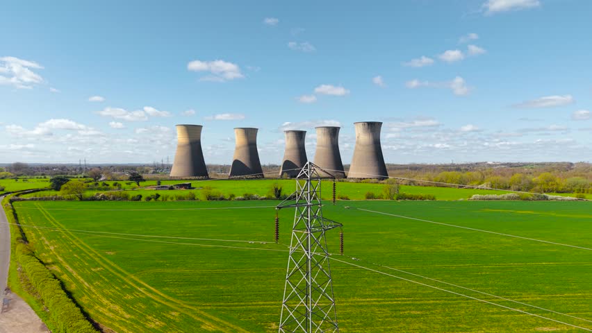 Wide view of power lines crossing green fields toward nuclear power station with multiple cooling towers. Energy infrastructure in rural landscape with transmission network and industrial background