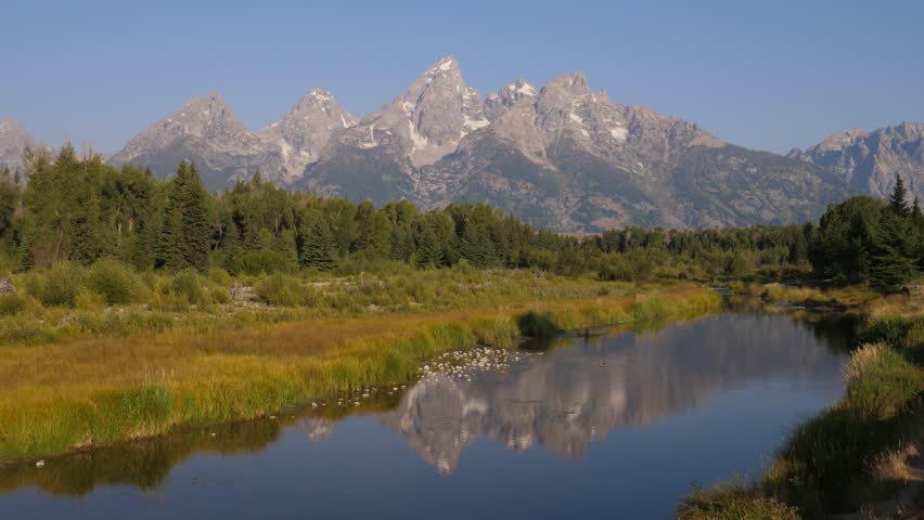 This 4K video shows the Teton Range in Grand Teton National Park, Wyoming. The visual documents the reflection of the Cathedral Group, including Grand Teton and Mount Owen, in a side channel of the Snake River at Schwabacher Landing. The composition illustrates the riparian habitat and calm water surfaces, documenting North American geography for nature and editorial media.