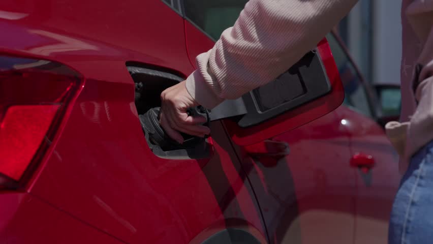 Close up of a female hand holding a green fuel pump nozzle and refueling a modern red car at a self service gas station on a sunny day