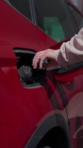 Close up of a hand unscrewing the fuel cap and inserting a green gas pump nozzle into a modern red car to begin refueling at a station
