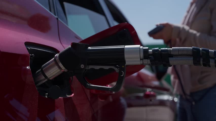Close up of a gas pump nozzle fueling a red car. In the background, a woman uses her smartphone while waiting for the tank to fill up