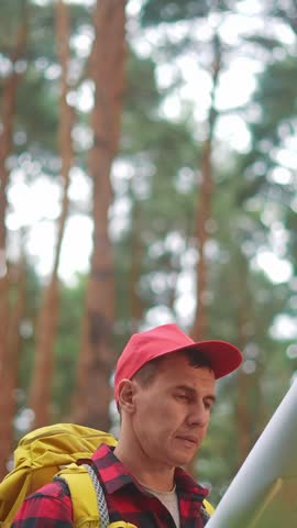 A man in a red cap studies a map while standing among trees. The man appears focused, navigating through the forest with the help of the map.
