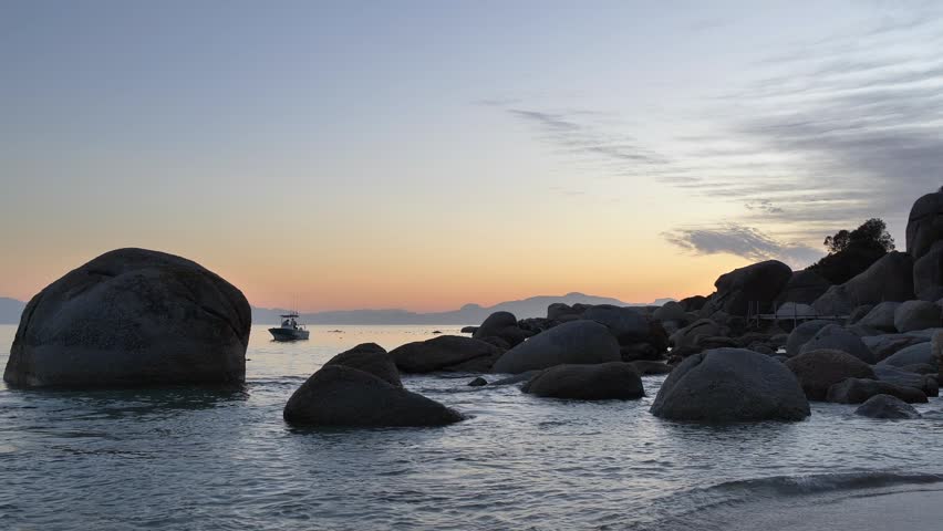 A tranquil dawn breaks over a rocky coastline, with a small boat gently moving through the serene ocean waters as the sun paints the sky with soft hues at Millers Point, Simon