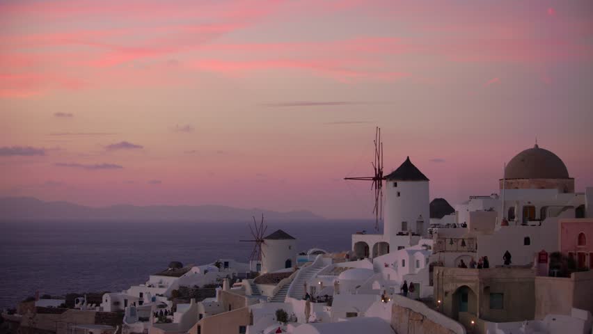 This 4K video shows the town of Oia on Santorini, Greece. The visual documents whitewashed settlements, blue-domed churches, and traditional windmills perched on caldera cliffs. The composition illustrates the Cycladic architecture and sunset lighting over the Aegean Sea, documenting Greek island geography and urban reconstruction for architecture, travel, and editorial media.