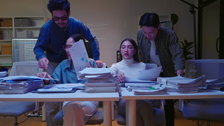 Group of Asian colleagues pan shot searching through disorganized piles of documents, feeling rushed and stressed to meet a night deadline.