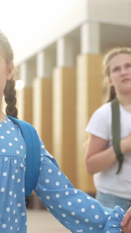 Two girls are holding hands in a school setting. One girl has curly hair while the other girl is wearing a blue shirt with polka dots.