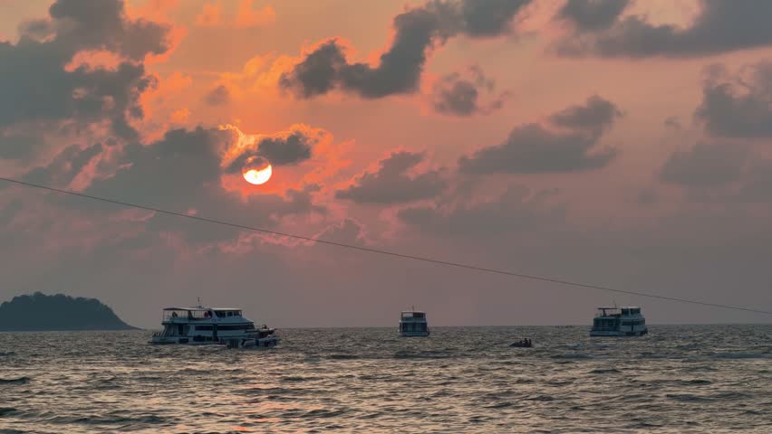 Parasailing activity over the sea at sunset on Phuket island, Thailand. Tourist flying with colorful parachute above the ocean with boats and jet skis on the shore during golden hour.