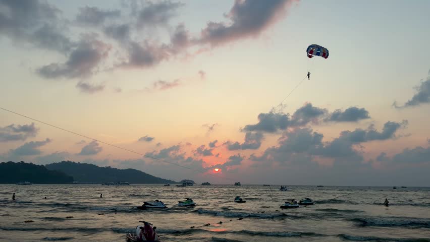 Parasailing activity over the sea at sunset on Phuket island, Thailand. Tourist flying with colorful parachute above the ocean with boats and jet skis on the shore during golden hour.