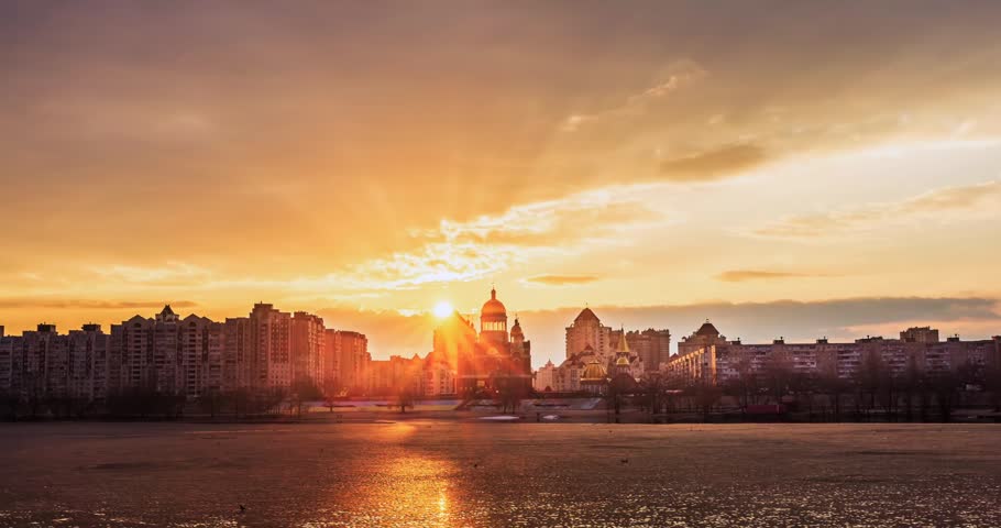 Soft sunset over Obolon embankment on the Dnipro River in Kyiv, Ukraine. Warm sky colors reflect on water, framing modern skyline and distant church in a calm urban evening scene.