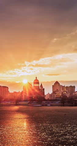 Soft sunset over Obolon embankment on the Dnipro River in Kyiv, Ukraine. Warm sky colors reflect on water, framing modern skyline and distant church in a calm urban evening scene.
