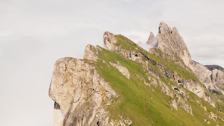 Aerial circular view Seceda ridge Odle peaks rise above clouds after rain with steep limestone cliffs green alpine slopes winding hiking trail panoramic Dolomites landscape South Tyrol Italy
