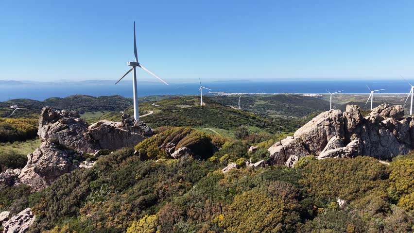 Low aerial pass over rocky outcrops and scrubland towards wind turbines on a ridge overlooking the Algeciras bay and the Strait of Gibraltar.
