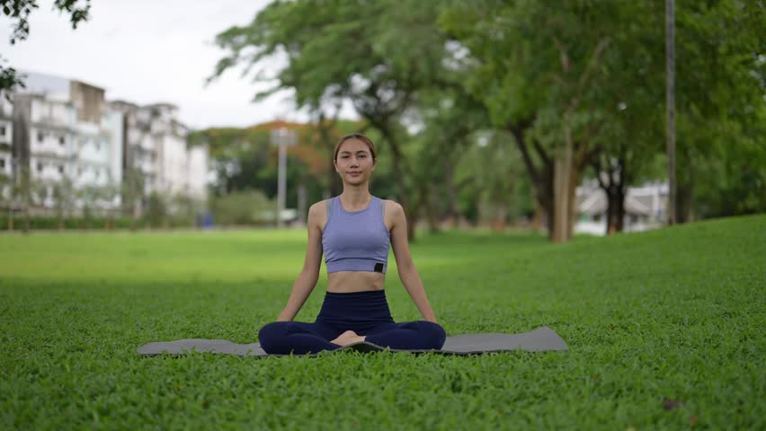 A woman is sitting on a mat in a park, looking at the camera. She is wearing a blue top and blue pants