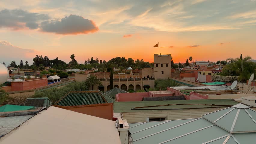 Panoramic view of Marrakesh medina rooftops at sunset with traditional architecture and orange sky. National flag of Morocco waving on top of the historic tower against the evening sky. Scenic skyline of the old city in Morocco featuring a historic stone tower and clay buildings. Tranquil evening atmosphere over the urban landscape with soft warm sunlight during golden hour. Wide shot of the moroccan city center capturing the authentic culture and rooftop terraces of the maghreb. Authentic urban landscape of old medina during evening time.