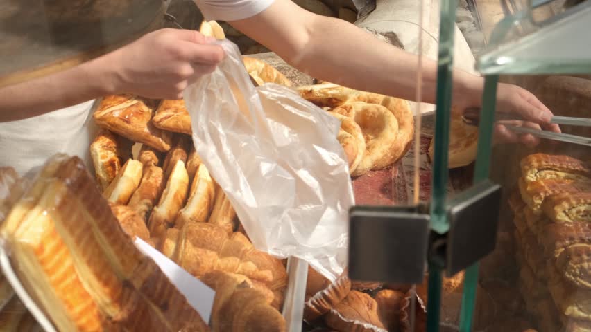 Baker uses metal tongs to pick and place fresh sweet traditional pastries into bag for customer. Pastry bakery filled with sunlight. Selling baked goods and bread. Bakery business