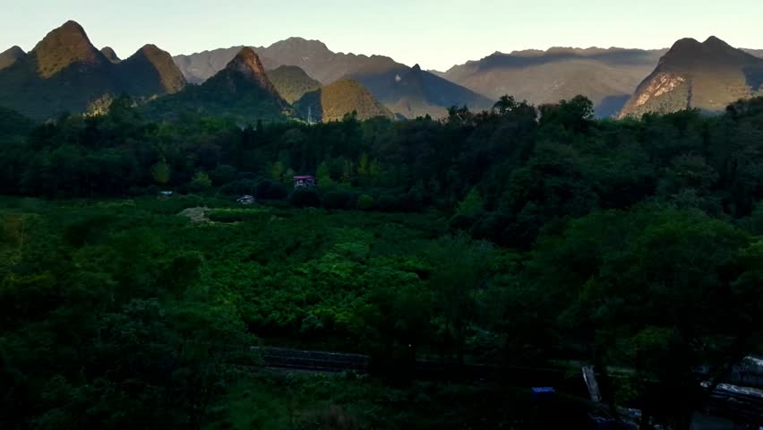 A serene landscape featuring lush green trees beneath a horizon of dramatic mountain peaks at dusk.